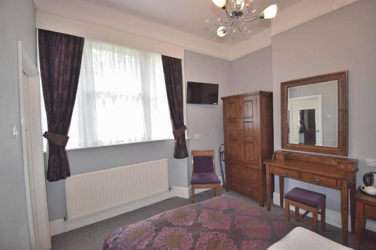 A bedroom with a bed, a wooden dresser, a chair, a mirror, and a television on the wall.