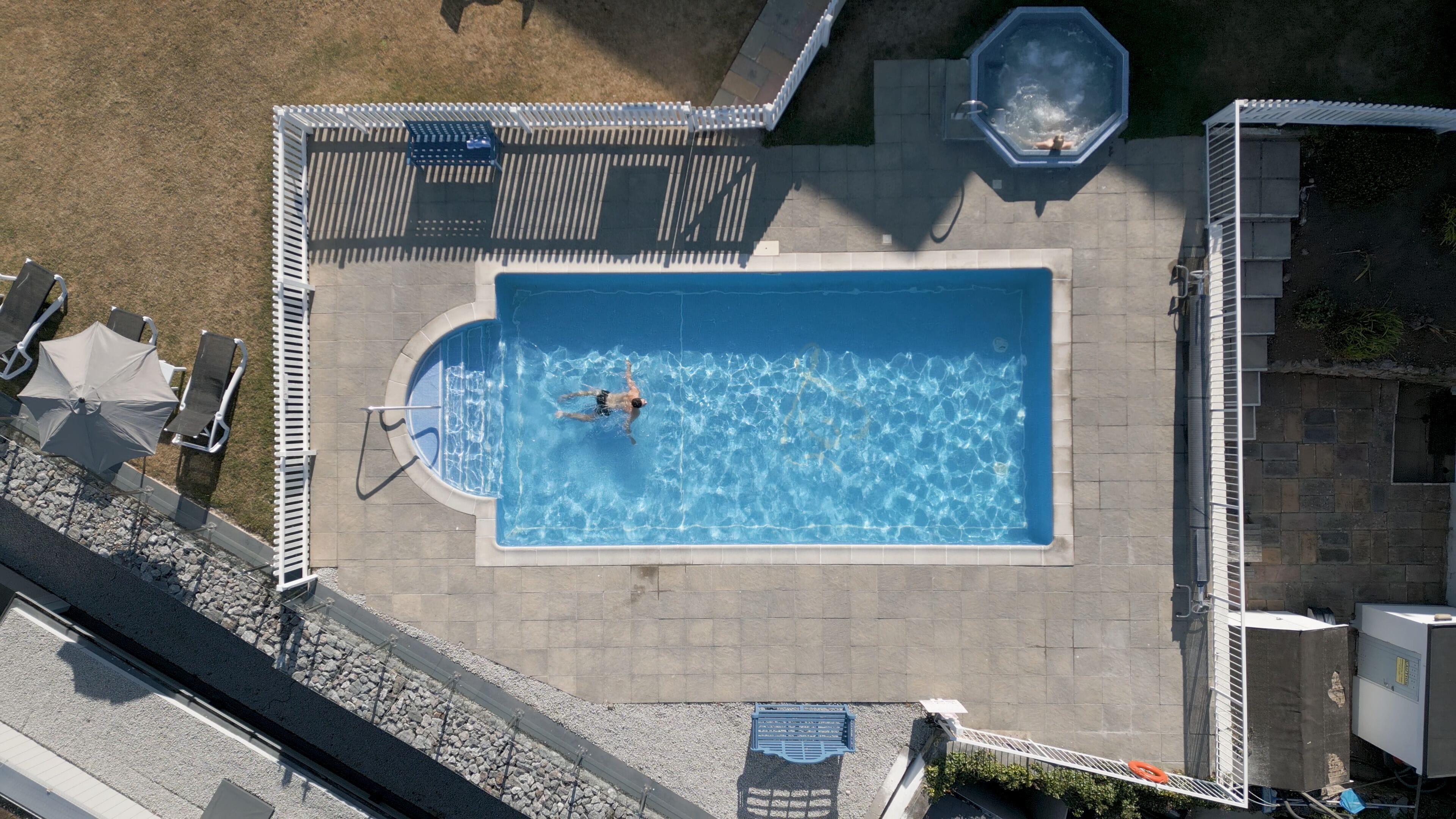 An aerial view of a rectangular swimming pool with a person swimming, surrounded by a deck area with chairs and umbrellas, and a hot tub to the right.