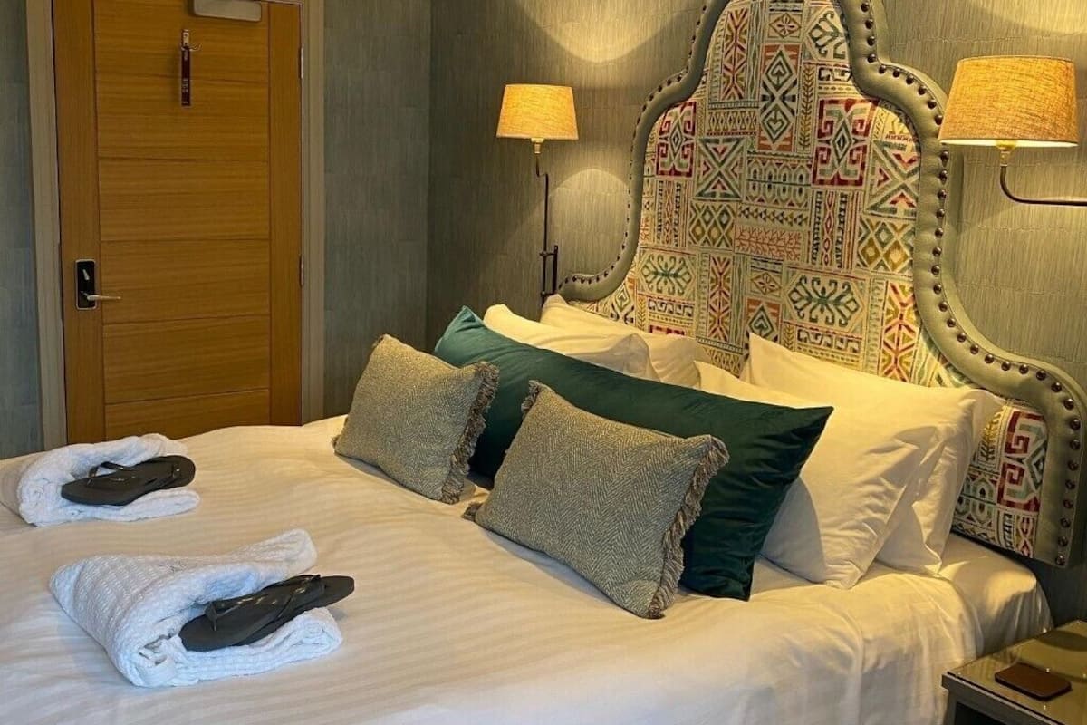 A neatly made bed with white linens and two towels, a wooden door, and a headboard with a patterned design.