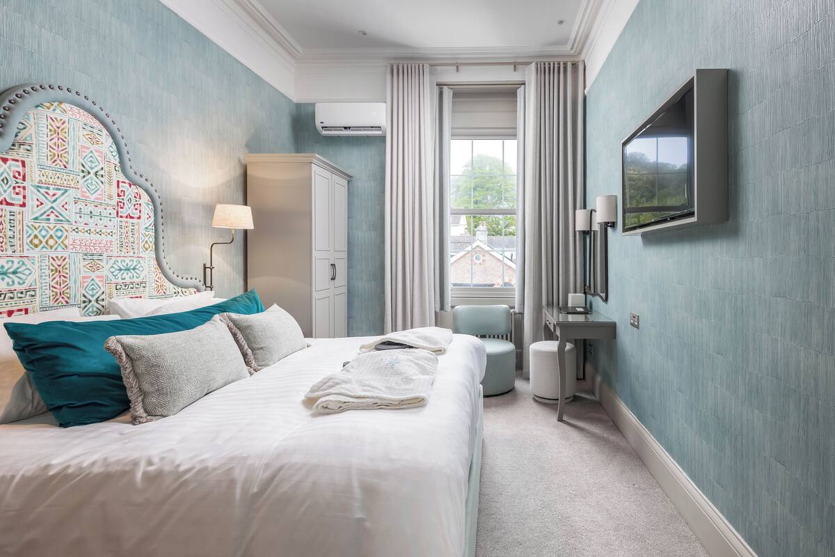 A bedroom with a large bed, a headboard with a colorful pattern, a television mounted on the wall, and a window with curtains.