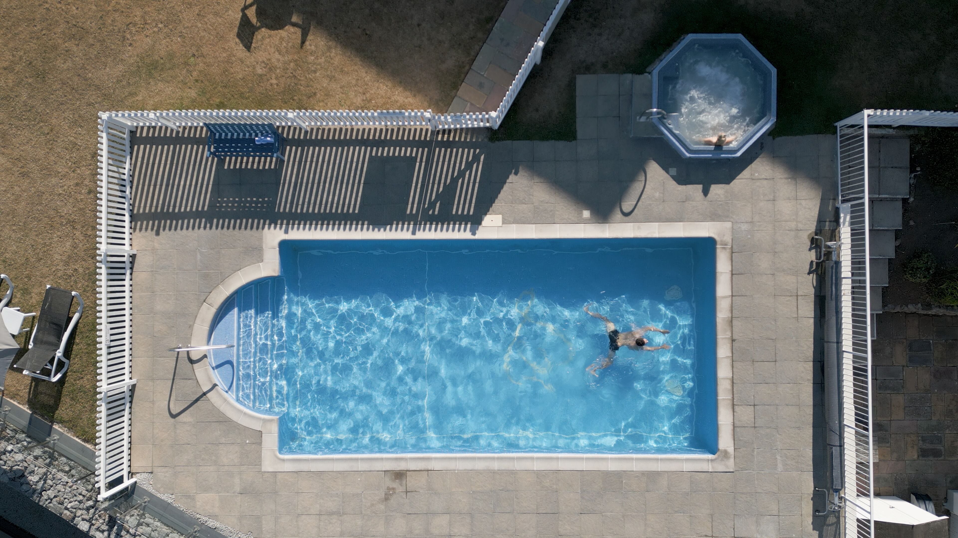 An aerial view of a rectangular swimming pool with a person swimming, surrounded by a tiled deck and a white fence.
