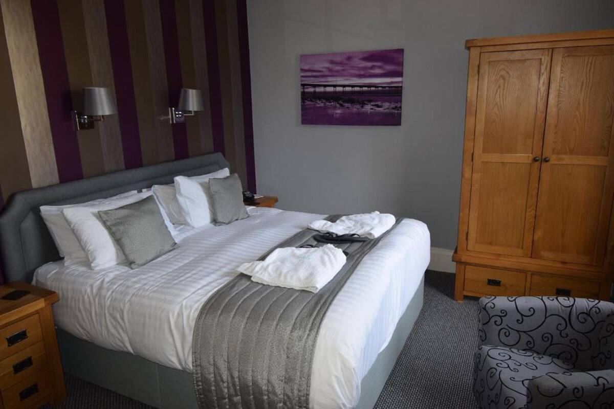 A neatly made bed with pillows and a blanket, a wooden wardrobe, a chair, and a wall-mounted lamp.