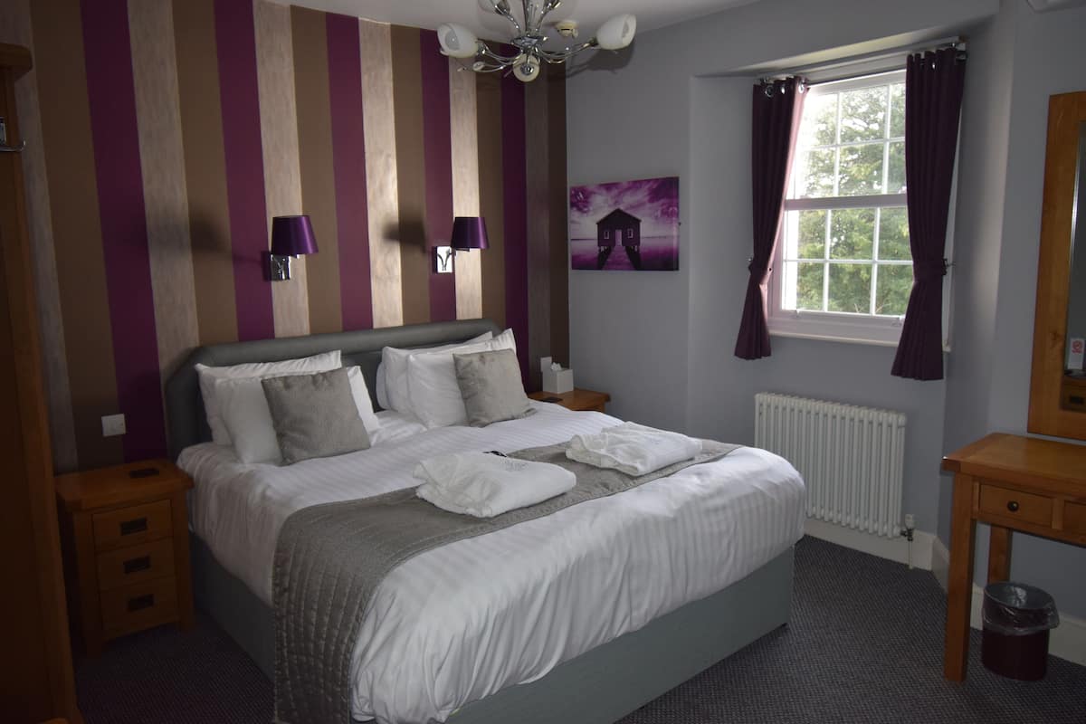 A neatly made bed with pillows and a blanket, a bedside table, a wall-mounted lamp, a window with curtains, and a radiator.