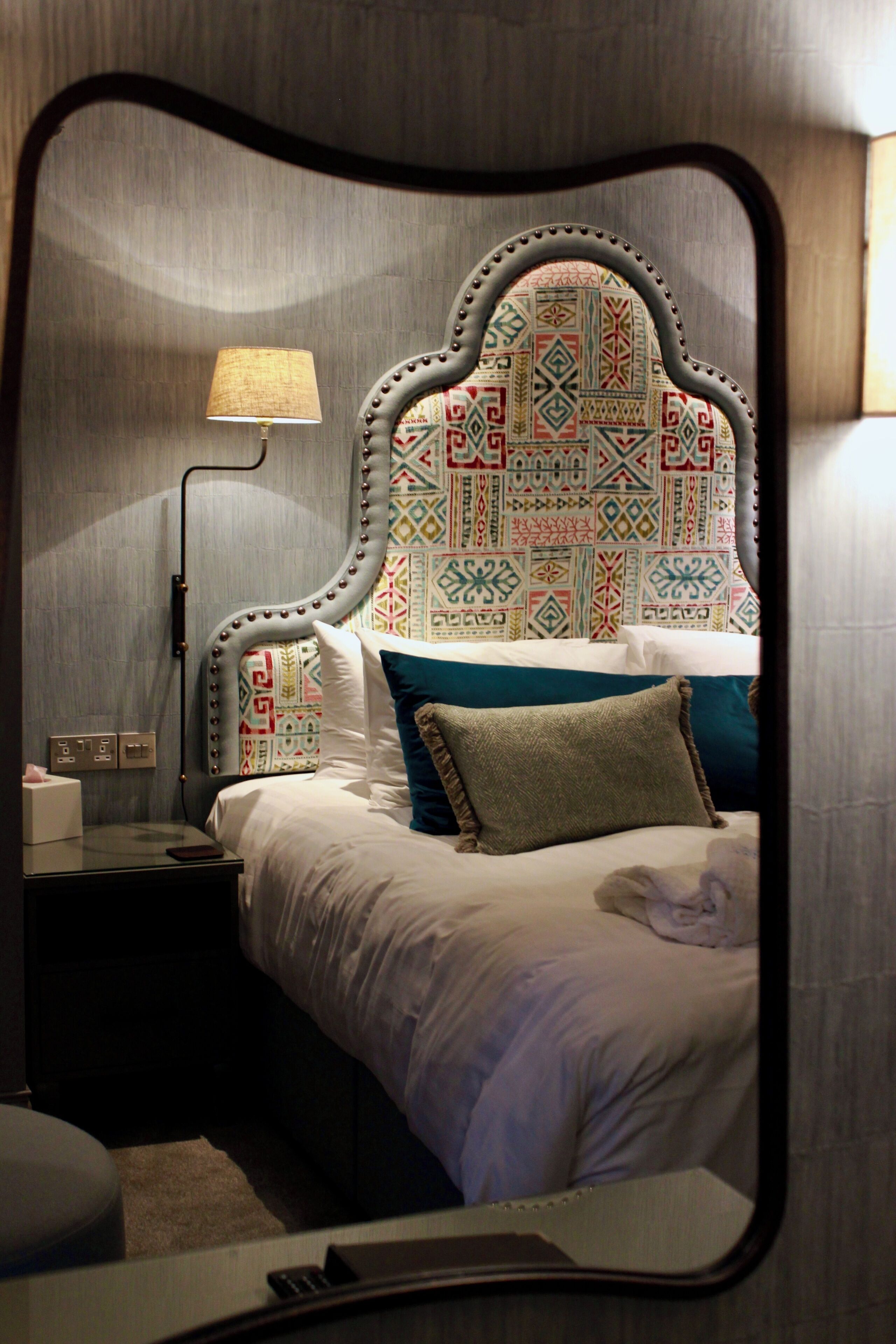 A hotel room with a bed, a nightstand, a lamp, and a mirror.