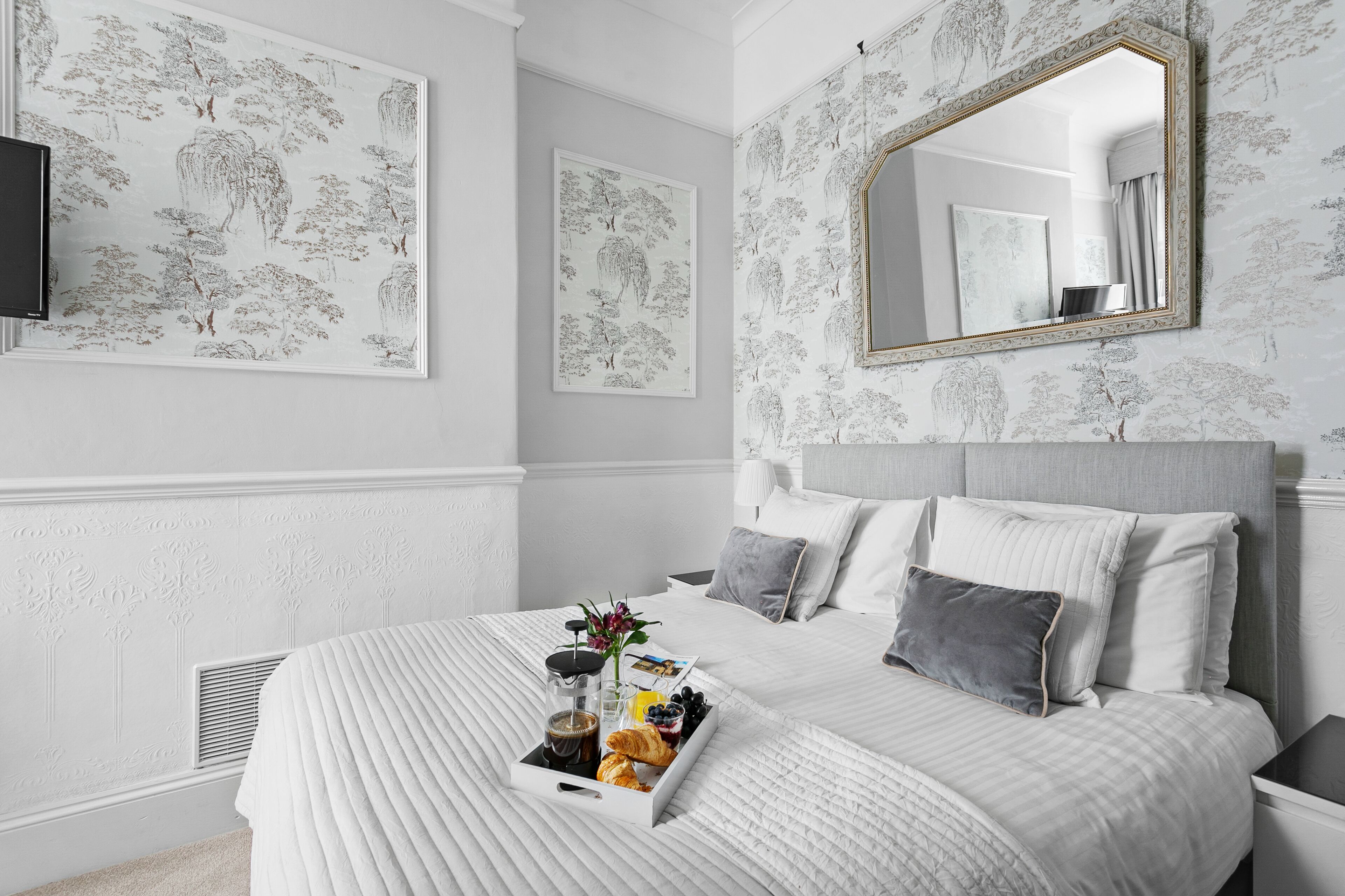 A neatly arranged bedroom with a bed, a bedside table, a mirror, and artwork on the walls.