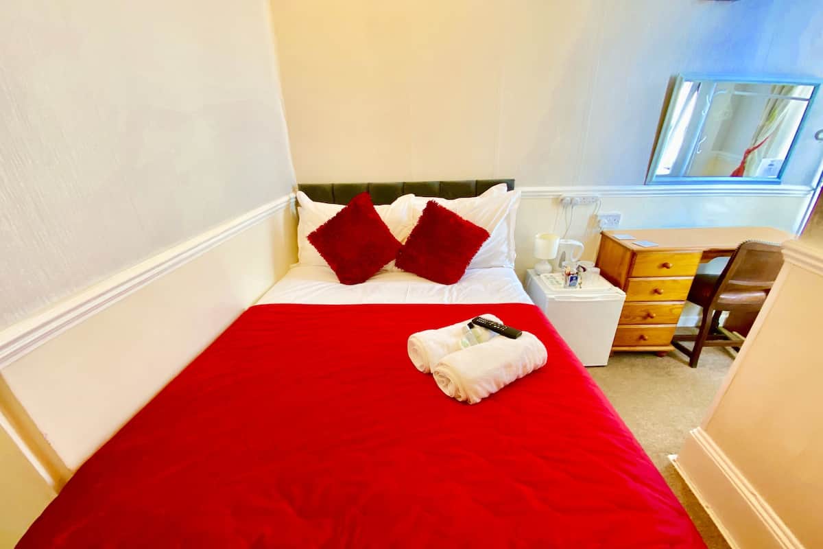 A hotel room with a red bedspread, white pillows, a white bedside table with a lamp, a wooden dresser, and a mirror on the wall.