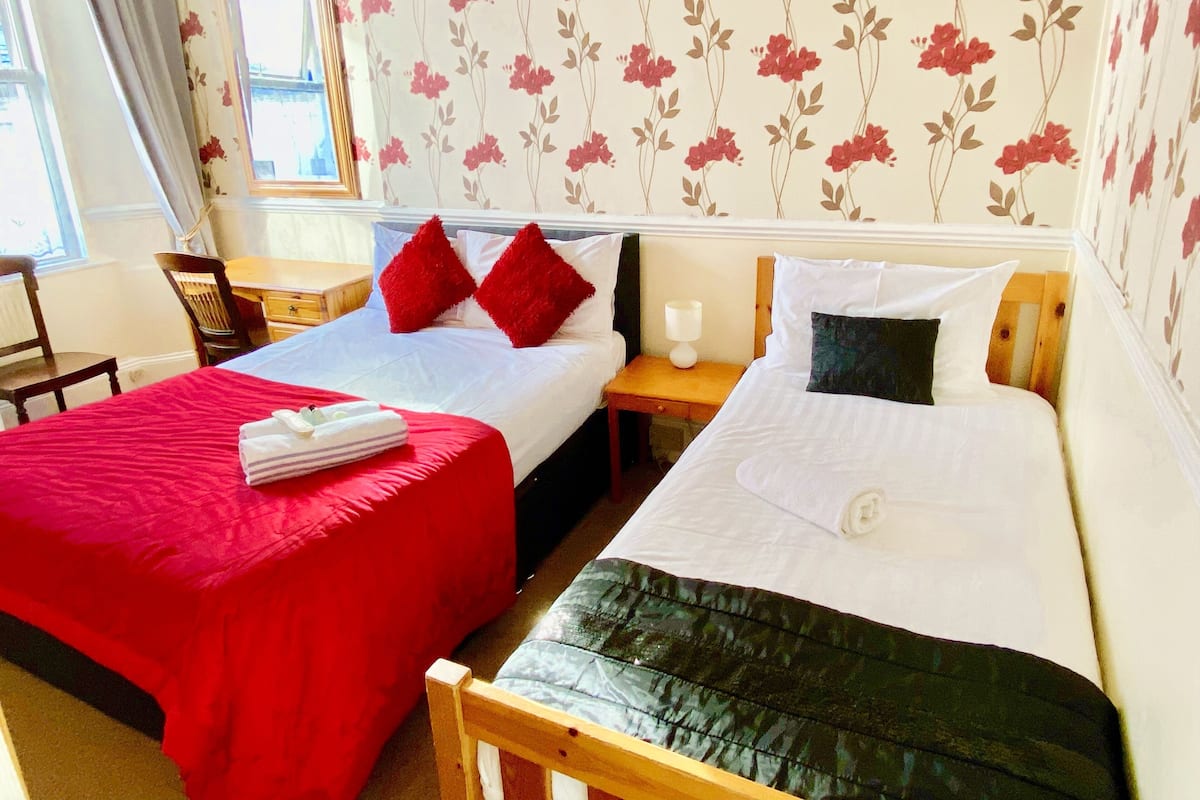 A hotel room with two beds, one with red bedding and the other with white bedding. There is a wooden nightstand with a lamp, a chair, and a window with curtains.