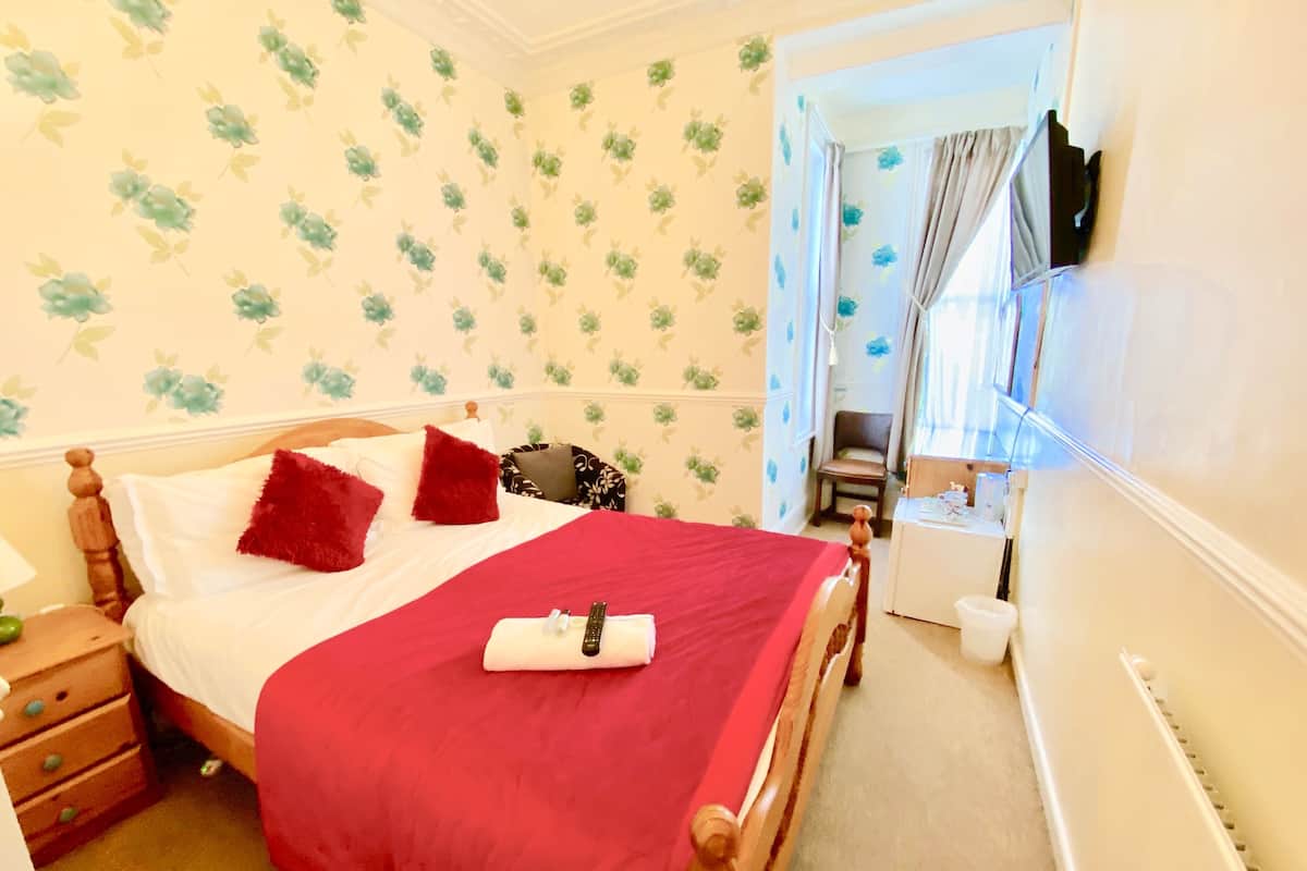 A bedroom with a wooden bed, red bedding, a small bedside table, a chair, and a window with curtains.
