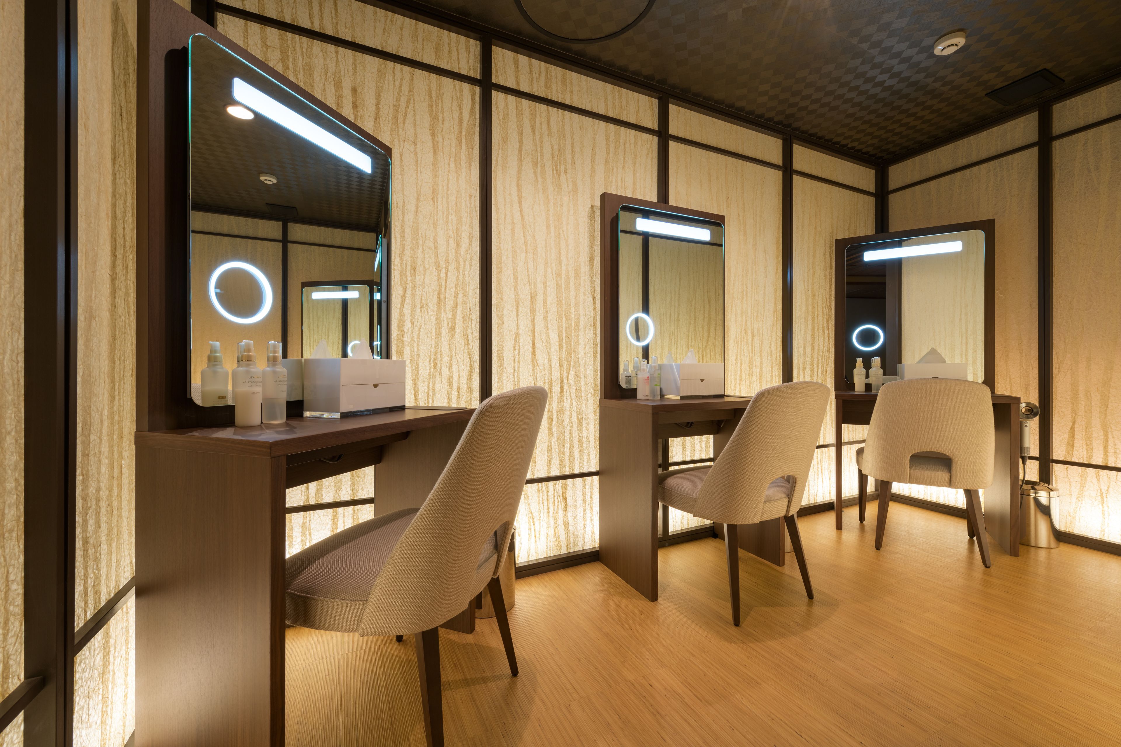 Una habitación con piso de madera, dos sillas, un escritorio con espejo y una tocador con espejo e iluminación.
