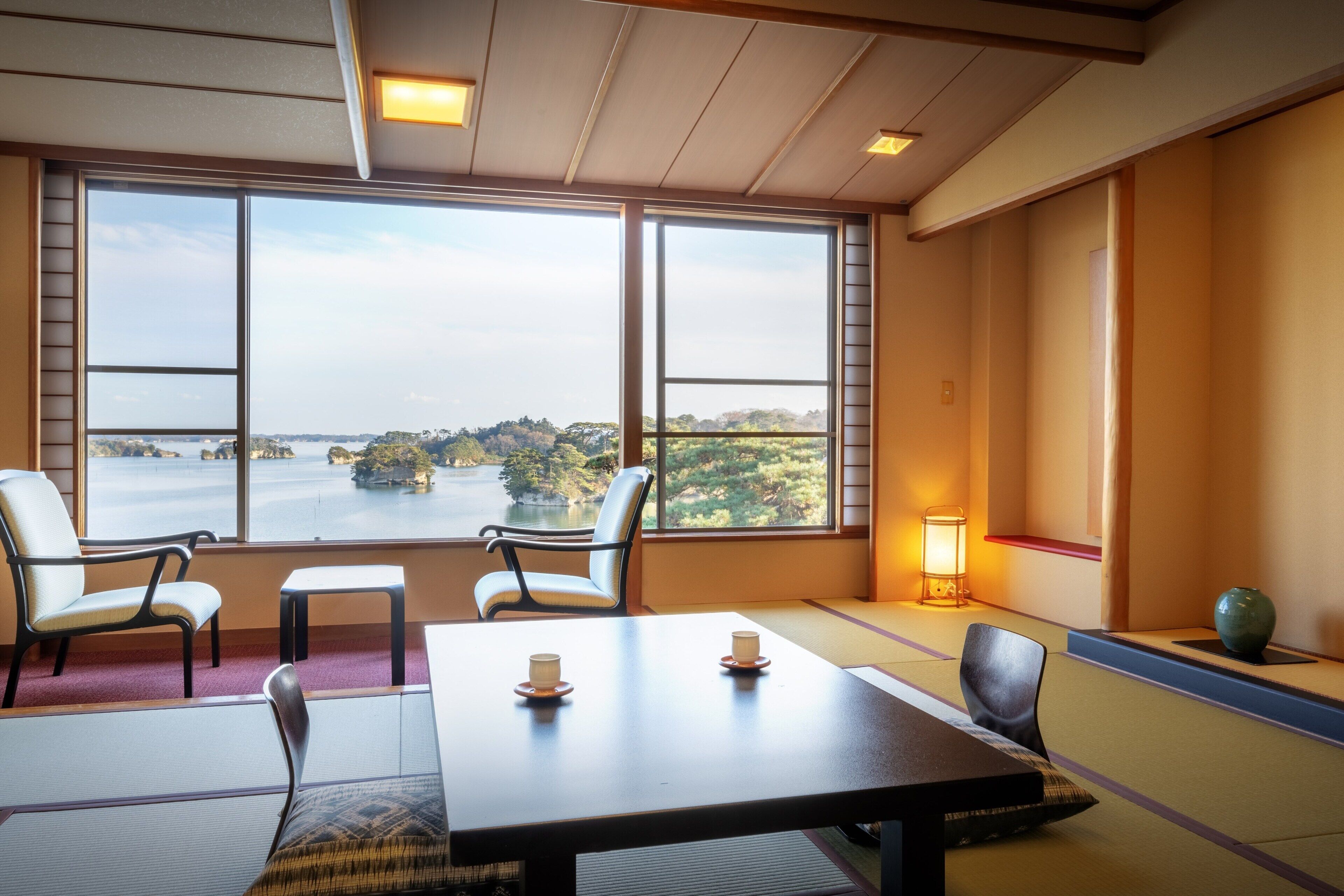 Una habitación con suelo de tatami, una mesa baja con dos tazas, una silla y vista a un cuerpo de agua.
