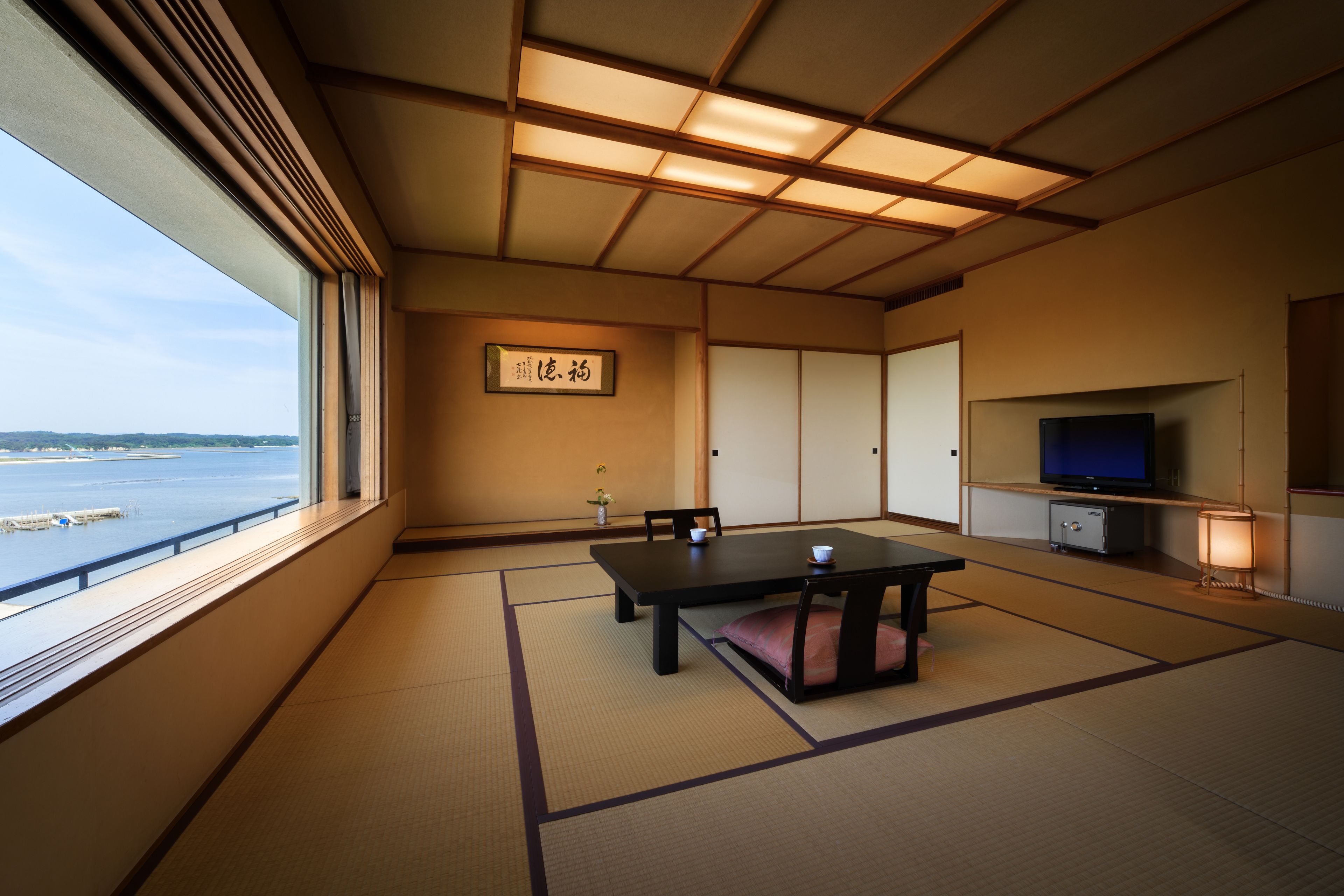 Une pièce de style japonais traditionnel, avec un sol recouvert de tatamis, une petite table basse avec des coussins, un téléviseur et une vue sur la mer.