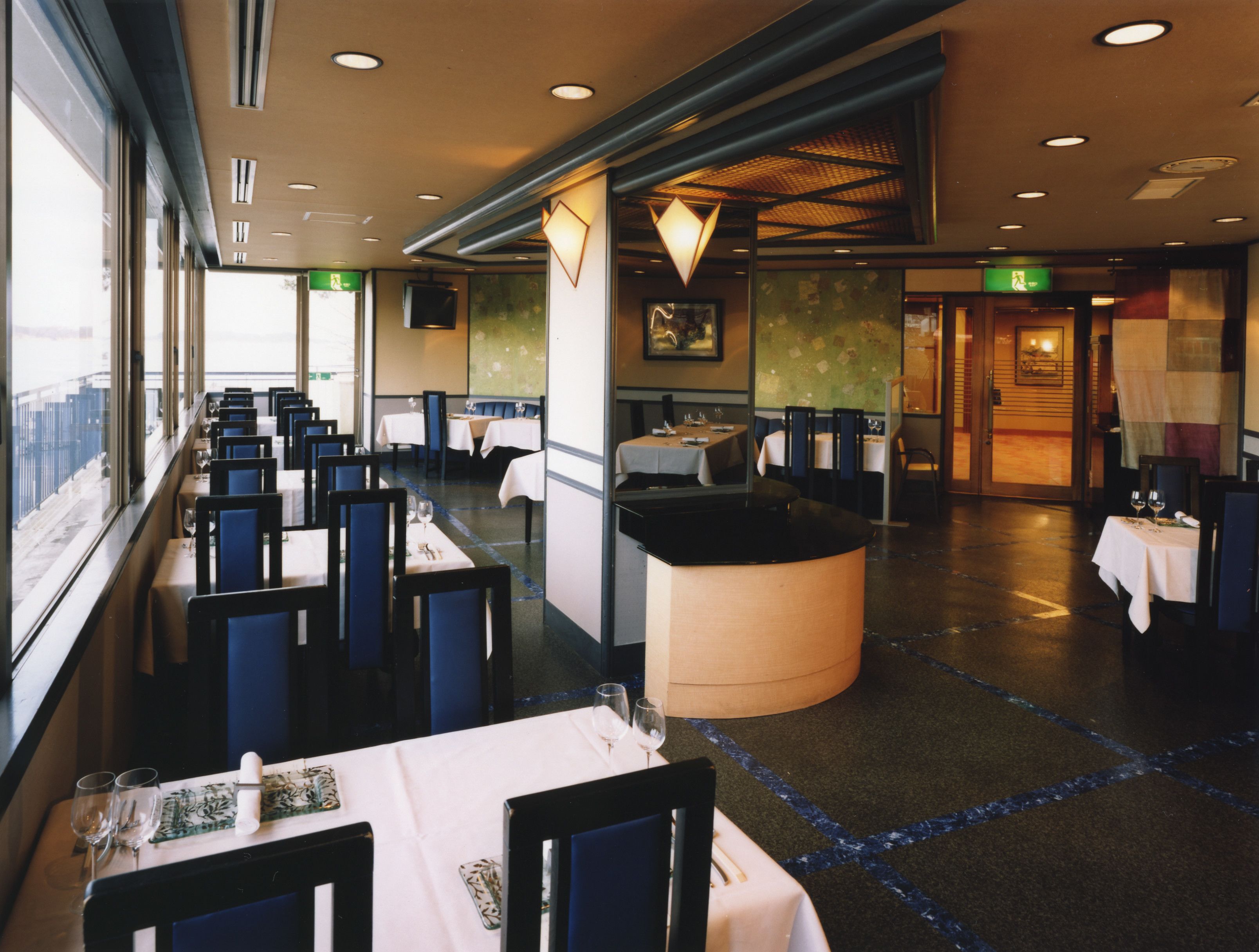 A restaurant with tables and chairs arranged in rows, a large window on the left, and a counter in the center.