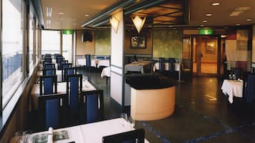 A restaurant with tables and chairs arranged in rows, a large window on the left, and a counter in the center.