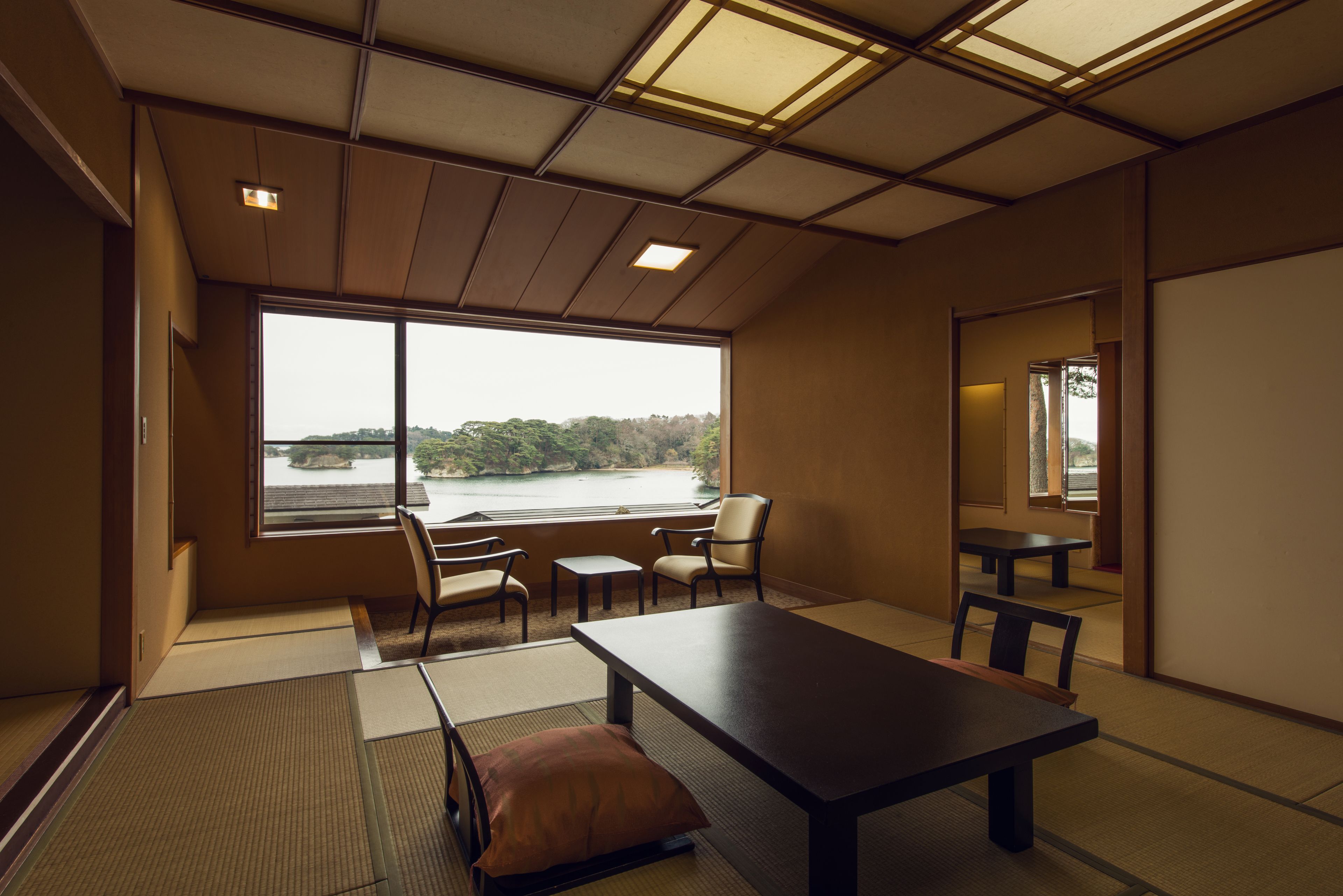 Une pièce de style japonais traditionnel, avec un sol recouvert de tatamis, une table basse, des chaises et une grande fenêtre donnant sur un plan d’eau.