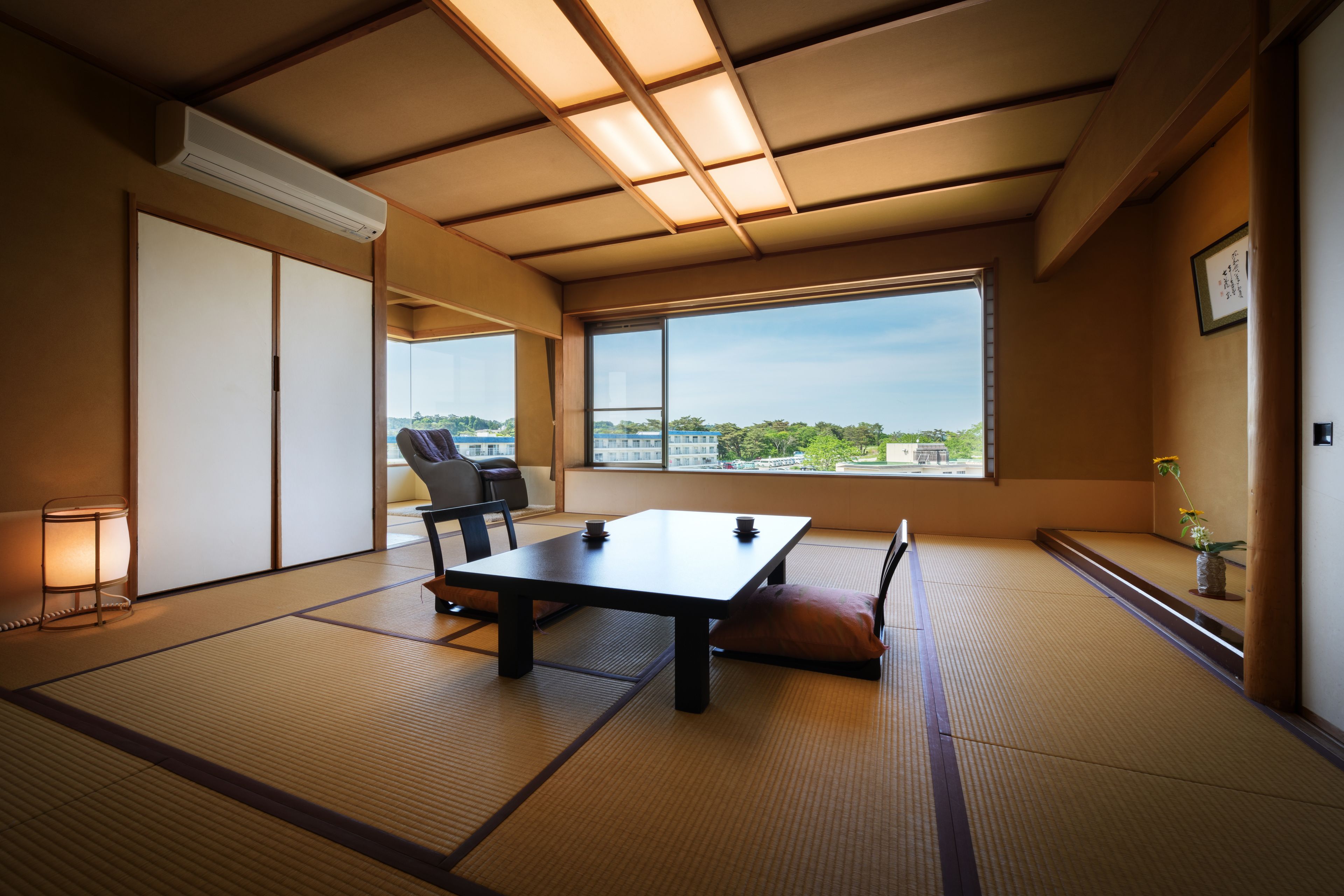A traditional Japanese room with tatami flooring, a low table, a chair, and a view of the city through large windows.