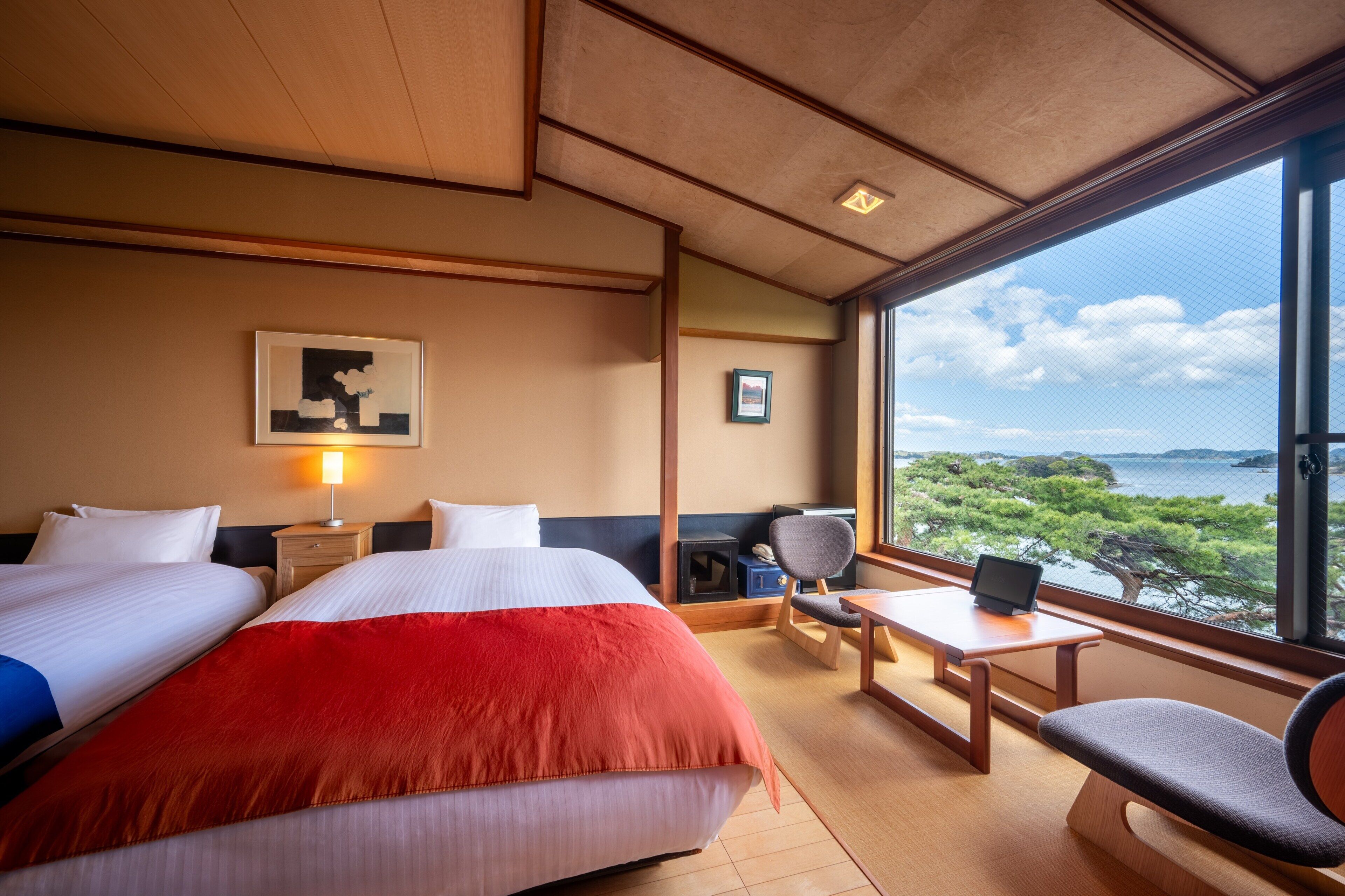A hotel room with two beds, a desk, a chair, and a large window offering a view of the sea.