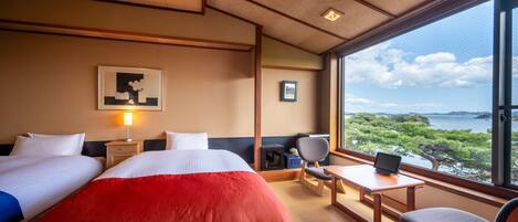 A hotel room with two beds, a desk, a chair, and a large window offering a view of the sea.
