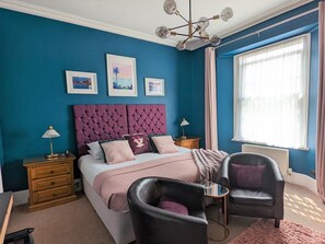 A bedroom with a tufted headboard, a bed with pink bedding, two nightstands, a black leather armchair, a small round table, and a window with curtains. - Villa Marina (Torquay)