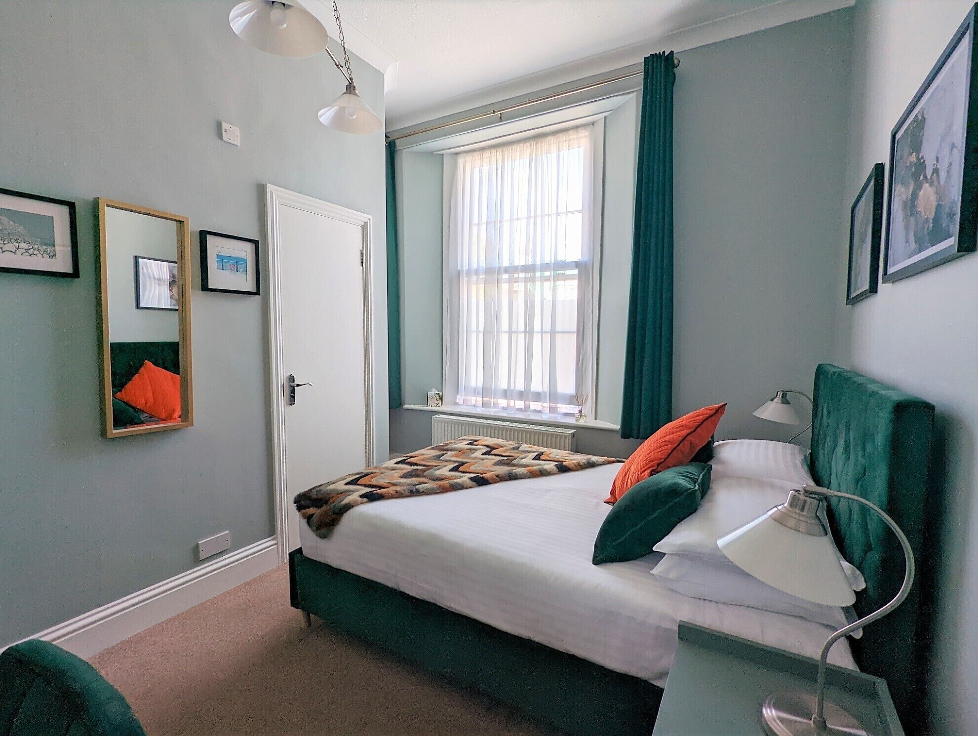 A bedroom with a bed, a bedside lamp, a window with curtains, a radiator, and framed pictures on the wall.