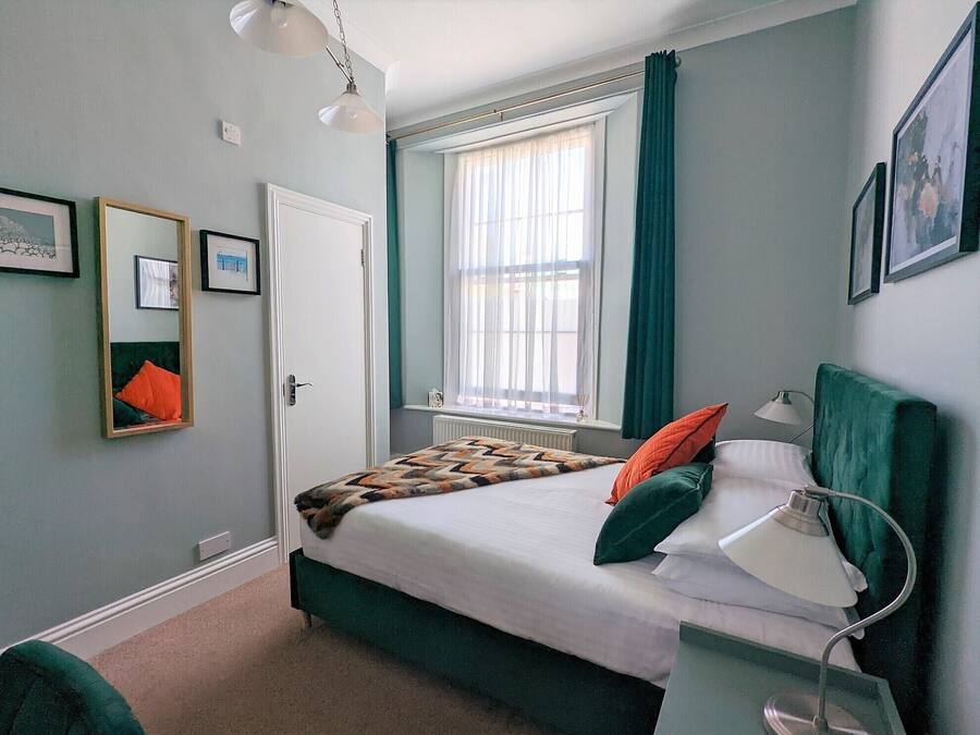 A bedroom with a bed, a bedside lamp, a window with curtains, a radiator, and framed pictures on the wall.
