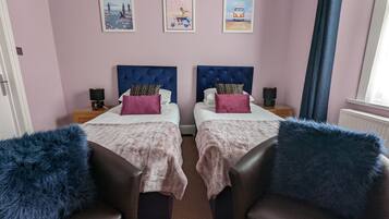 Habitación doble con dos camas, cada una con cabecero azul, y un par de sillas adornadas con mantas de pelo azul. Hay tres carteles enmarcados en la pared.