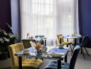 A dining area with tables set for two, featuring glass tops and upholstered chairs in yellow and blue. - Villa Marina (Torquay)