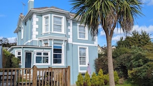 A two-story house with a bay window, a wooden deck, and a large palm tree in the front yard. - Villa Marina (Torquay)