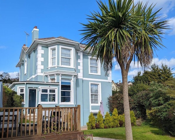 A two-story house with a bay window, a wooden deck, and a large palm tree in the front yard. - Villa Marina (Torquay)
