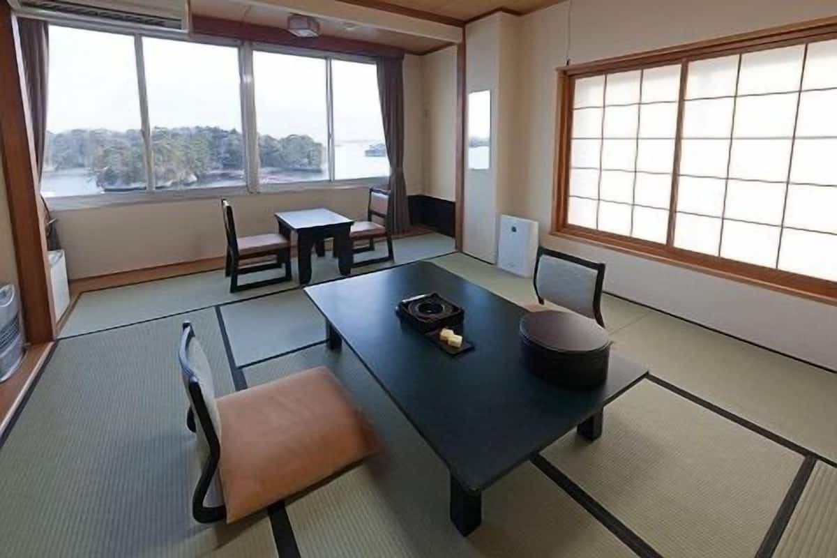 A traditional Japanese room with tatami flooring, a low table, chairs, and large windows offering a view of trees and water.