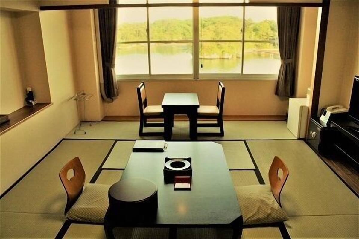 Ein traditioneller japanischer Raum mit Tatami-Bodenbelag, einem niedrigen Tisch mit Kissen und Blick auf eine grüne Landschaft durch große Fenster.