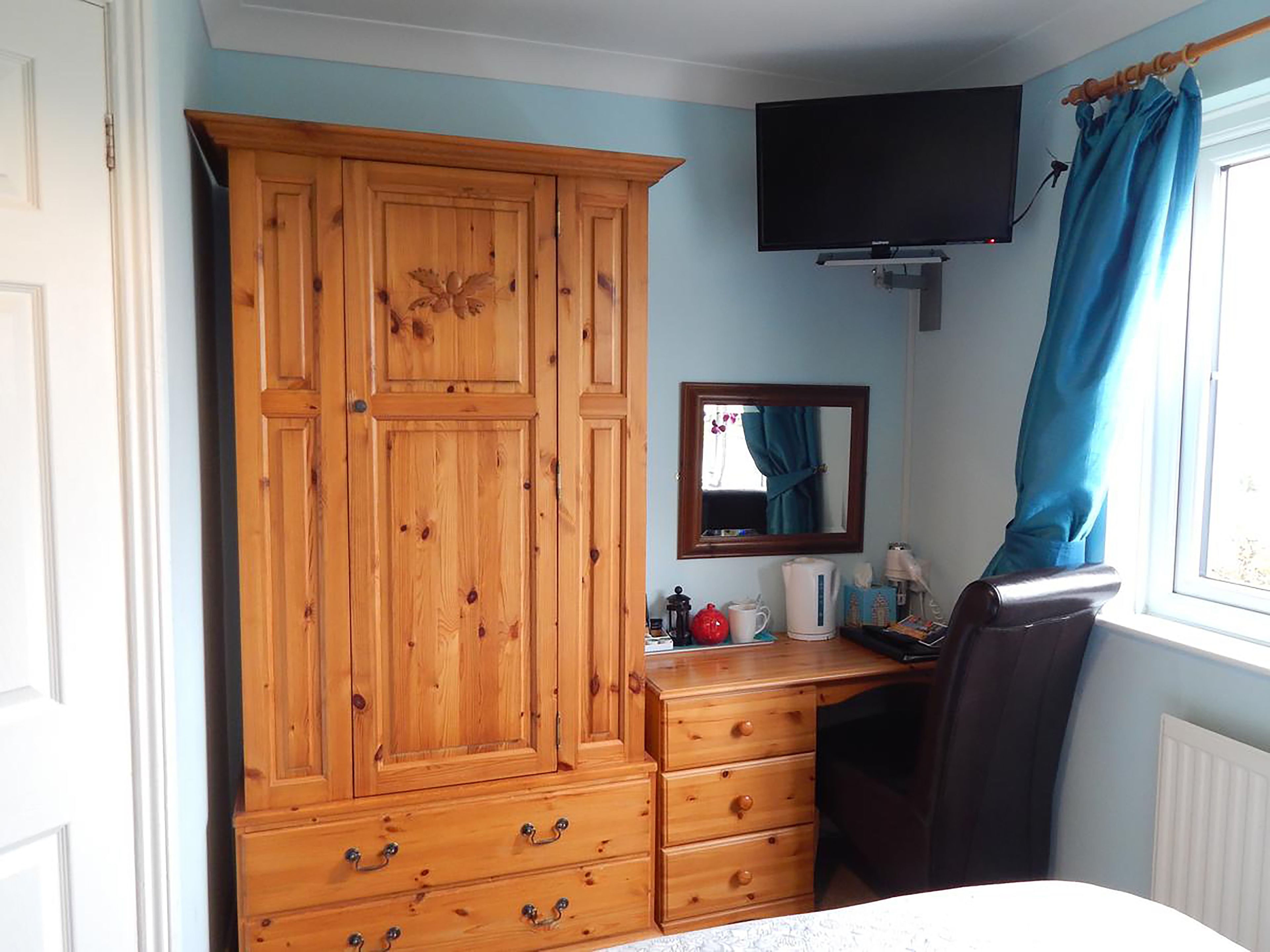 Een slaapkamer met een houten garderobe, een bureau met spiegel, een televisie en een raam met gordijnen.