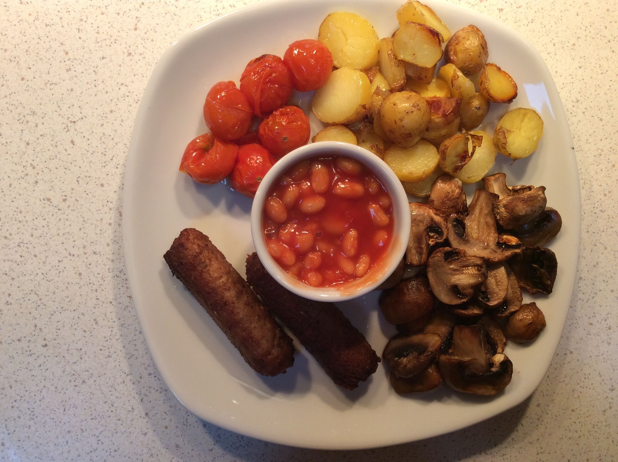 Um prato com batatas assadas, tomates cereja, cogumelos, feijão cozido e salsichas.