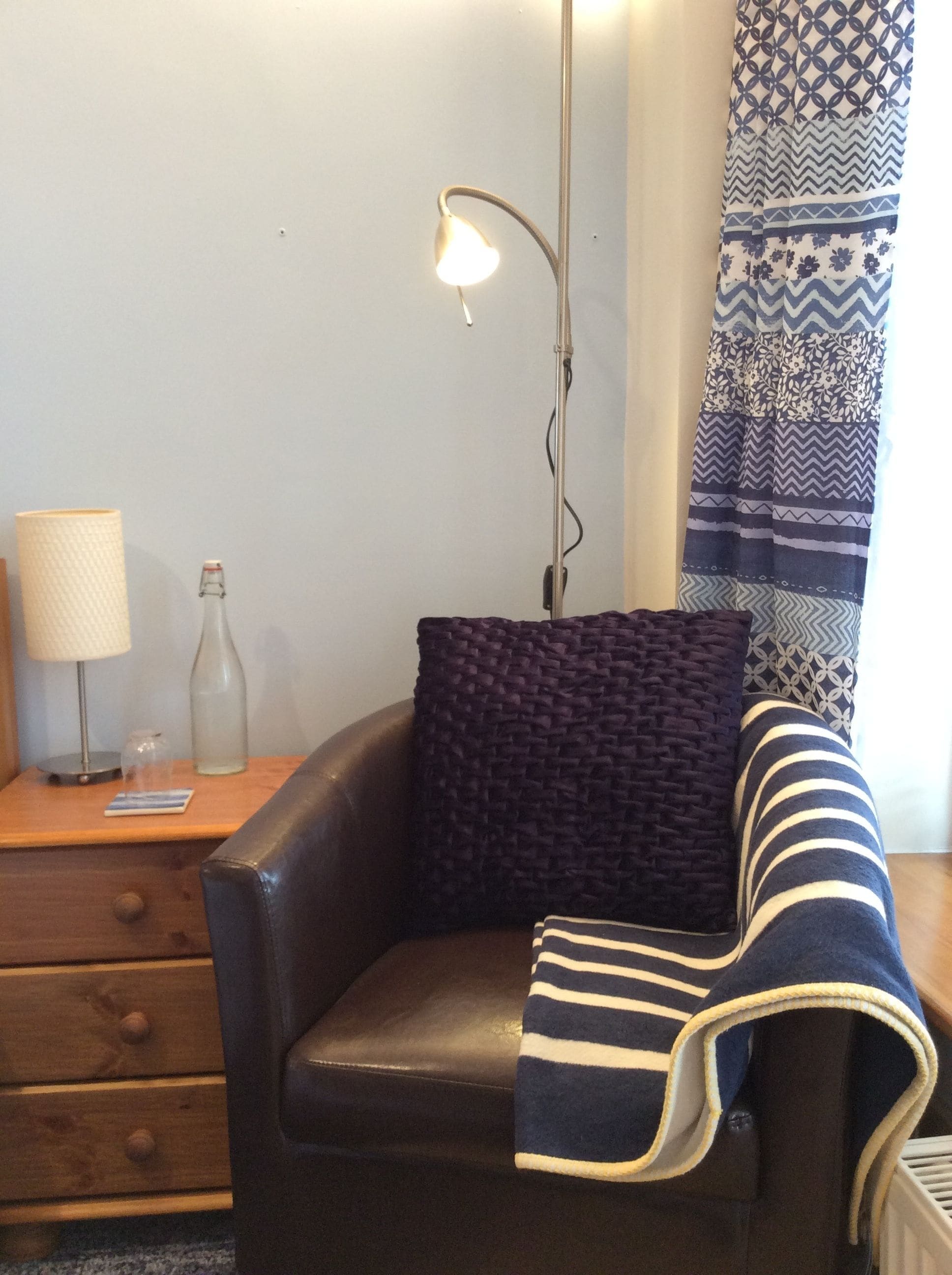 A cozy corner with a leather armchair, a patterned throw blanket, a lamp, and a bottle on a wooden side table.
