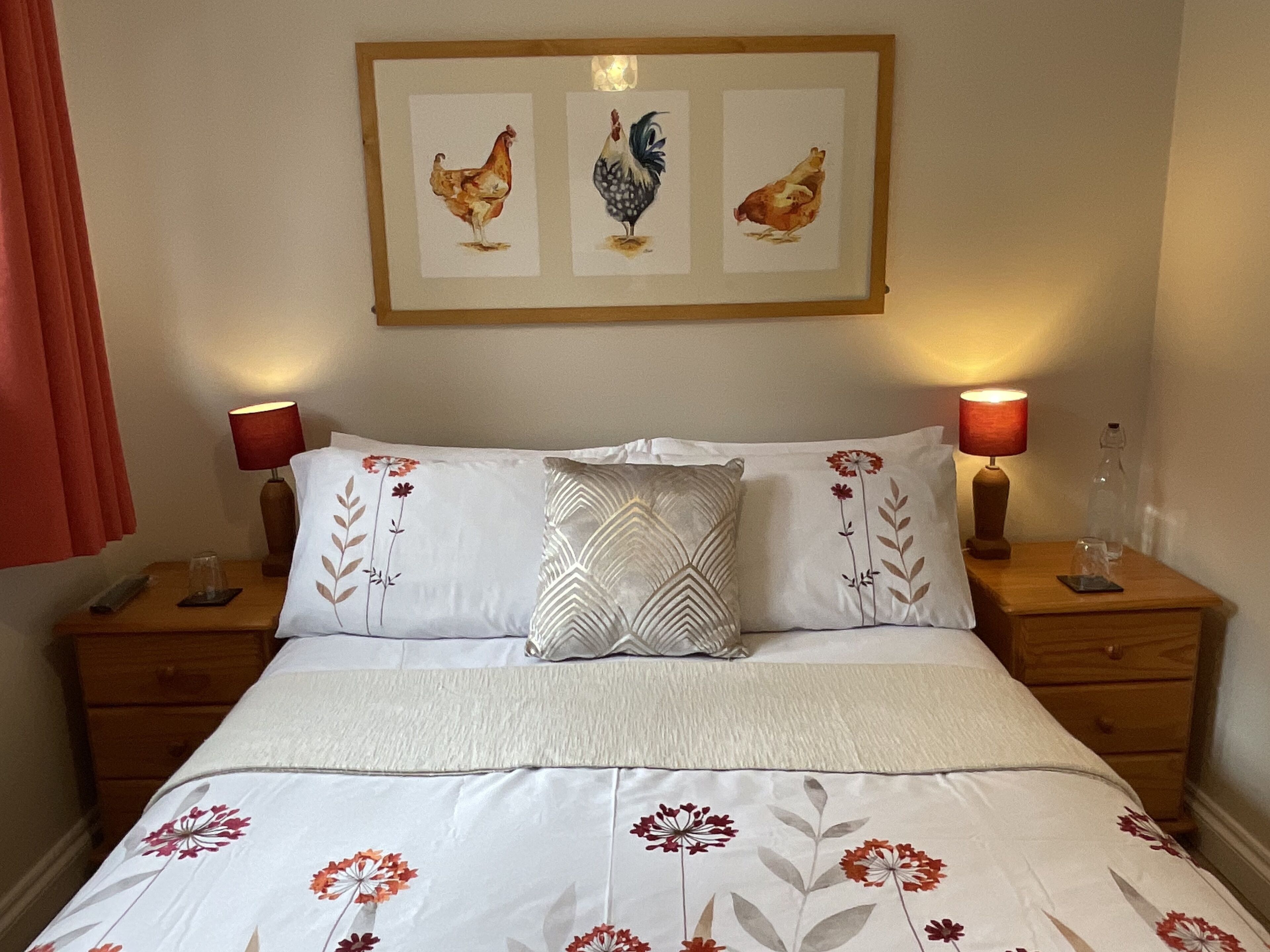 Un letto rifatto con lenzuola a motivi floreali, due abat-jour, un quadro appeso raffigurante galline e un comodino in legno con un telecomando.