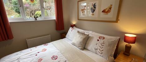 A bedroom with a bed, a window with curtains, a radiator, a bedside table with a lamp, and wall art.