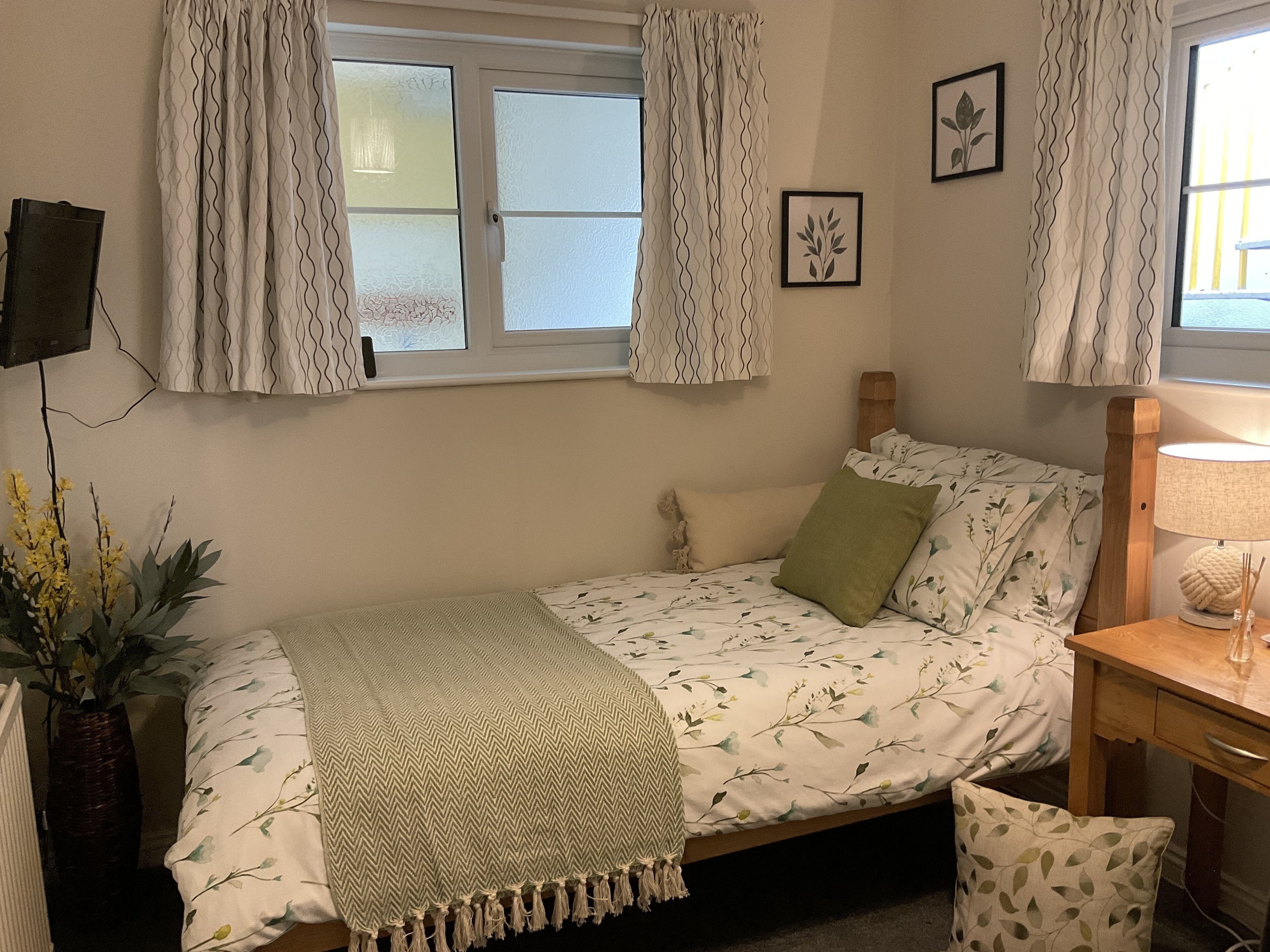 A bedroom with a bed, a bedside table with a lamp, a window with curtains, a wall-mounted TV, and a framed picture.
