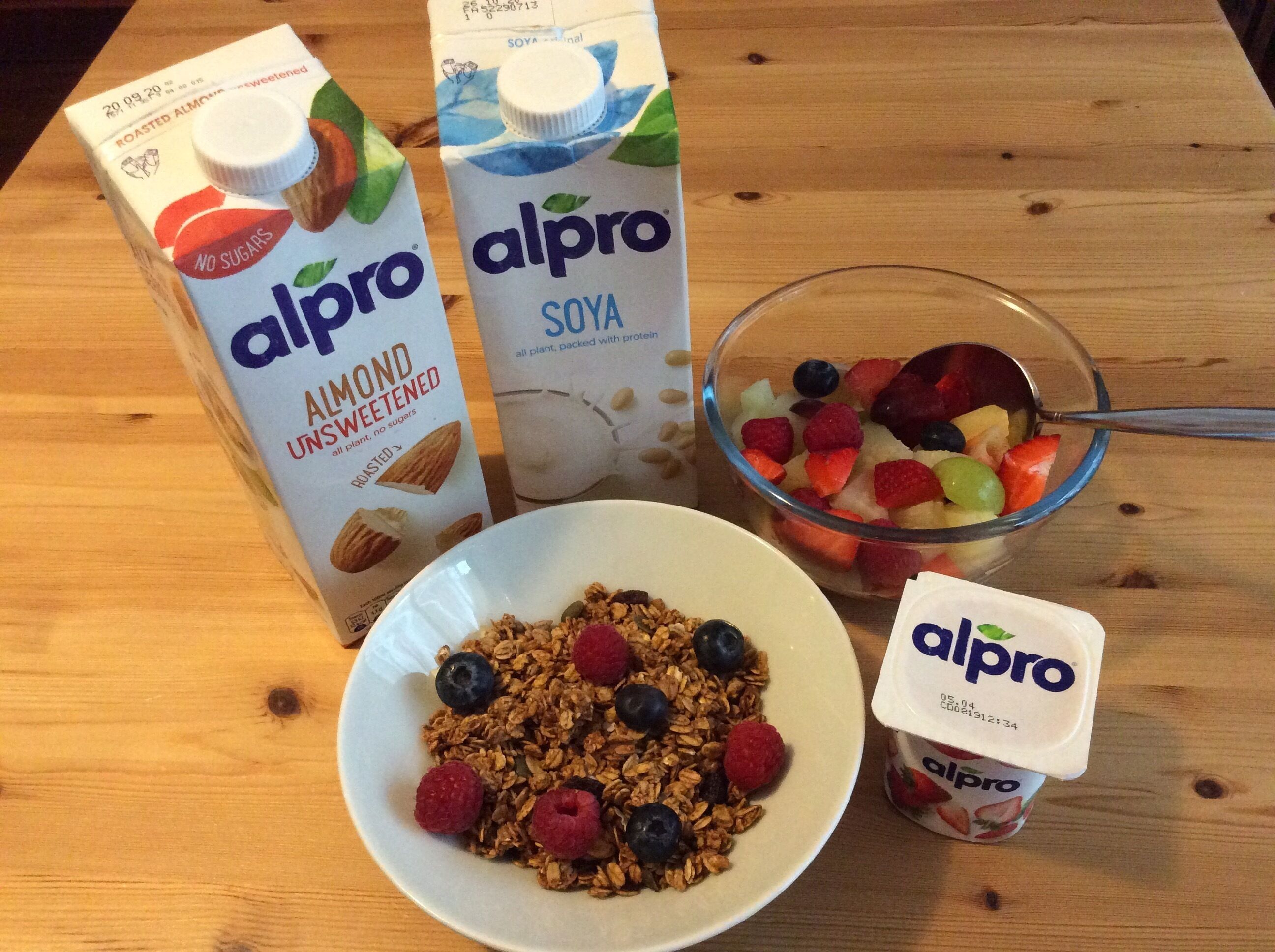 A breakfast setting with a bowl of granola, fresh berries, a glass of soya milk, and two cartons of almond unsweetened soya milk.