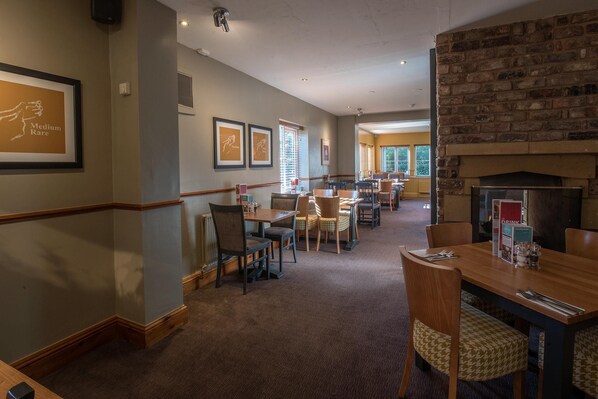 A hotel dining area with multiple tables and chairs, framed artwork on the walls, and a brick fireplace. - Premier Inn Walsall - M6 J10 (Walsall)