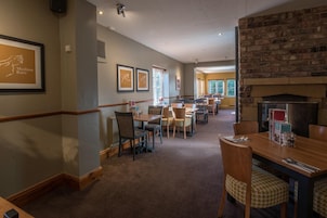 A hotel dining area with multiple tables and chairs, framed artwork on the walls, and a brick fireplace.