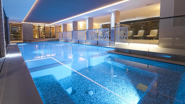 Un espace piscine intérieur moderne, avec une eau d’un bleu limpide, un coin salon avec des fauteuils et une paroi en verre.