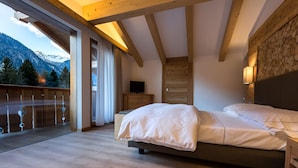 Une chambre avec un plafond en bois, un lit avec une literie blanche, un téléviseur posé sur une commode en bois et un balcon offrant une vue sur les montagnes.