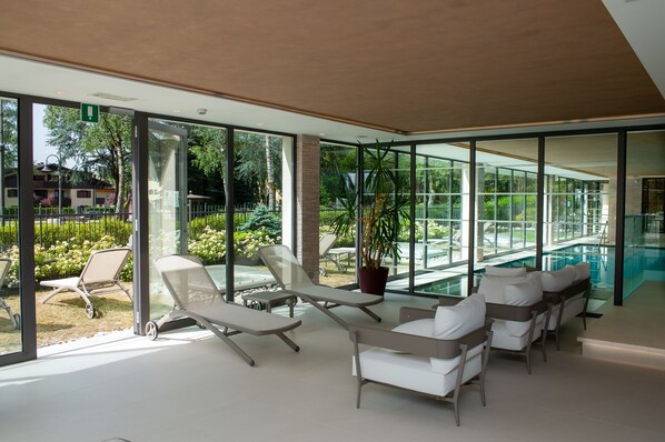 Uma área interna de piscina moderna, com paredes de vidro, espreguiçadeiras e um vaso de planta.