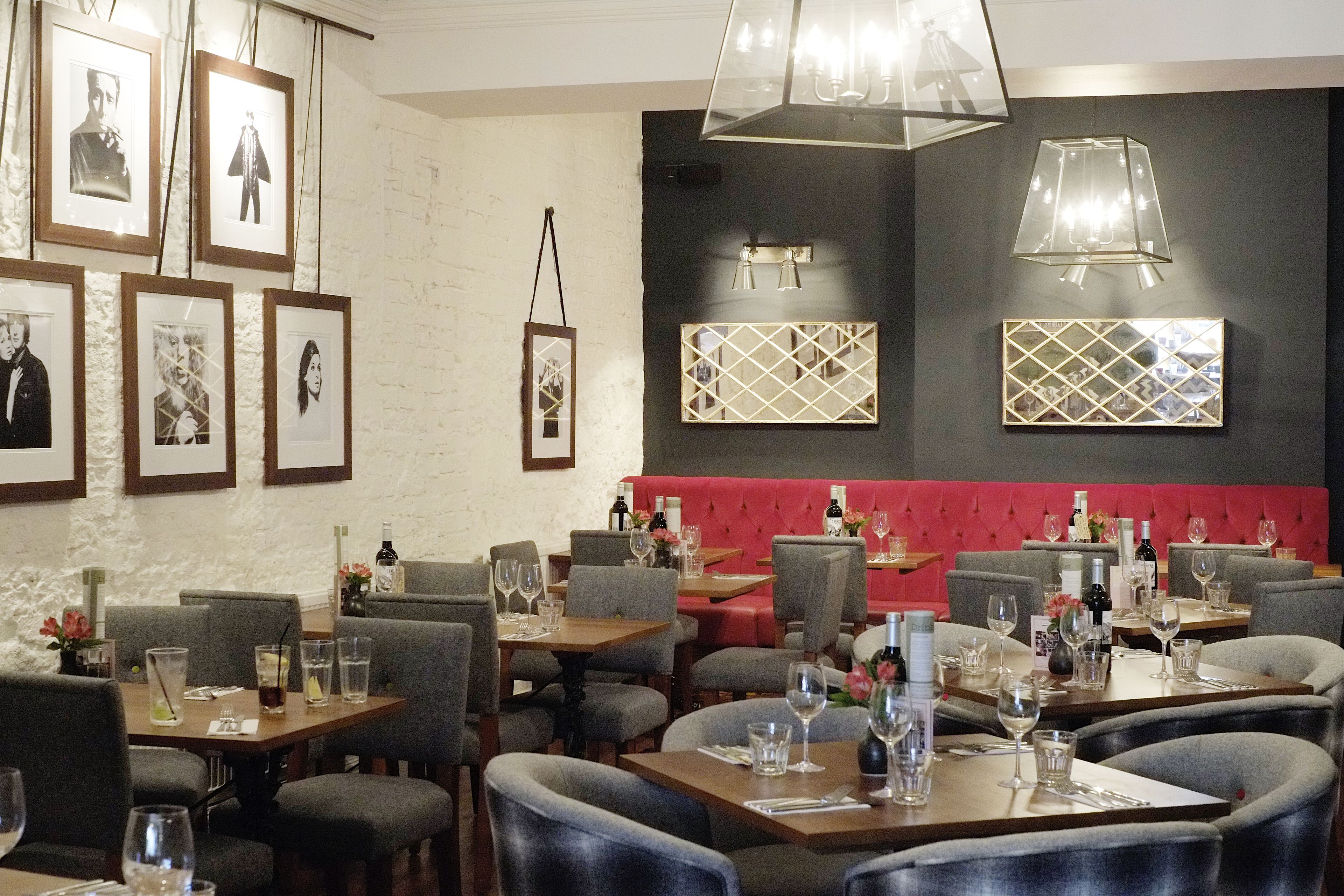 A restaurant with tables set for dining, framed pictures on the wall, and a red cushioned bench area.
