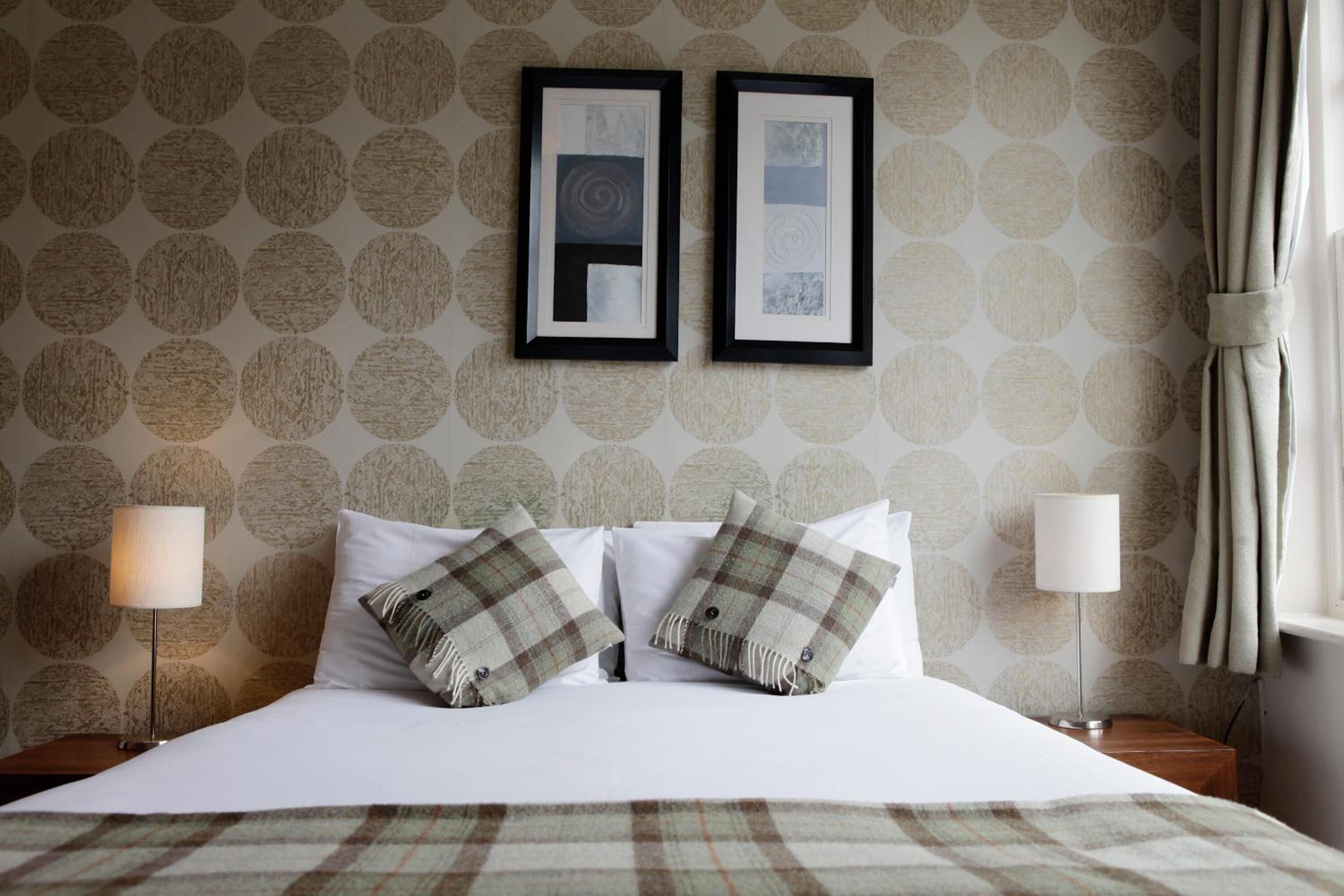 A neatly made bed with plaid bedding, two framed artworks on the wall, and a lamp on either side.