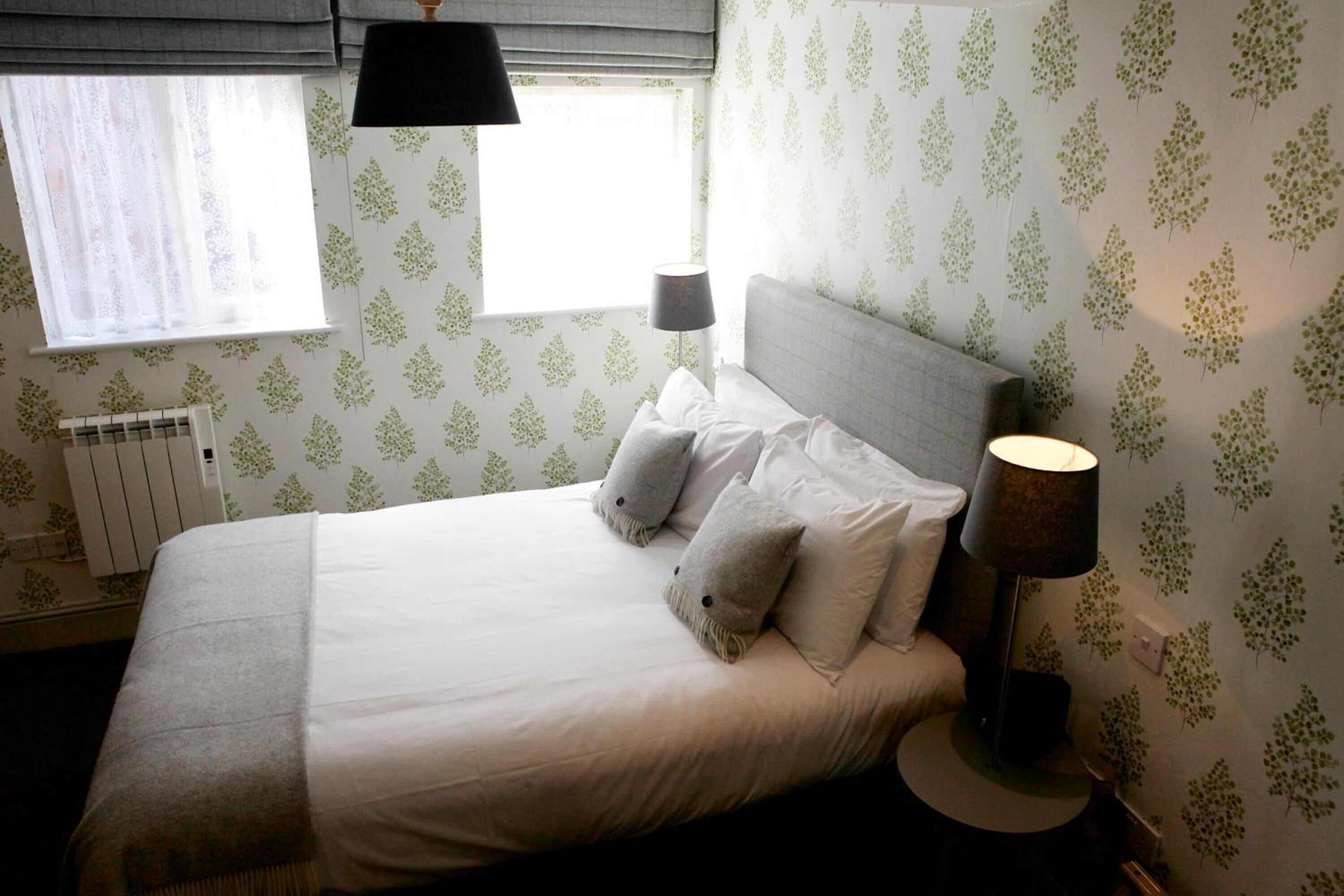 A neatly made bed with white linens and two pillows, a bedside lamp, and a window with sheer curtains.