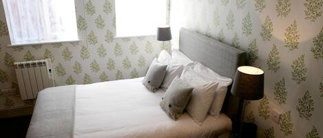 A neatly made bed with white linens and two pillows, a bedside lamp, and a window with sheer curtains.