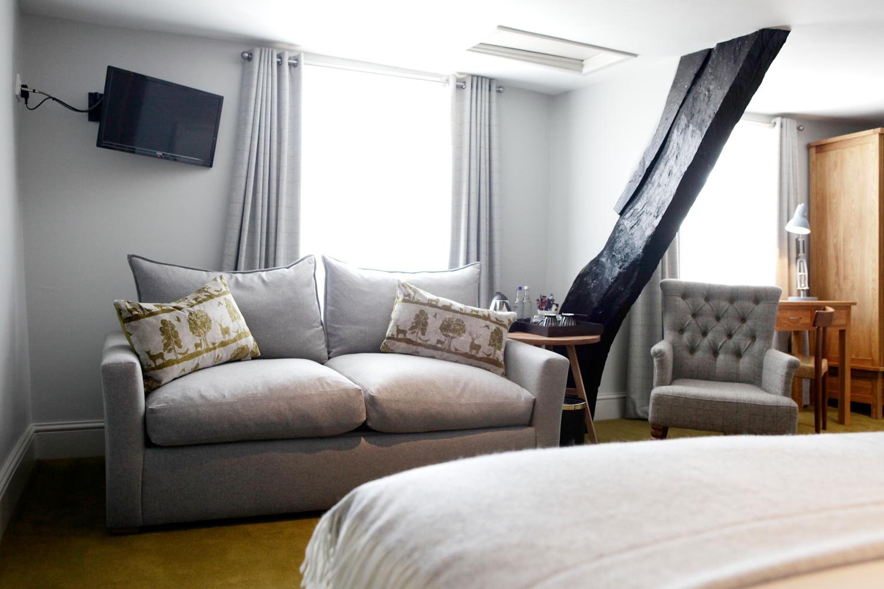Una habitación de hotel moderna con un sofá gris, un sillón pequeño, una cama, un televisor montado en la pared y un mueble de madera.