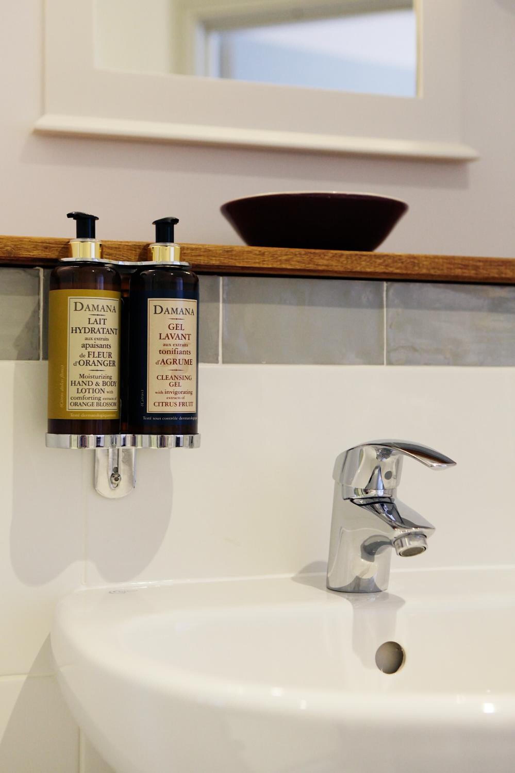 Baño con un lavamanos blanco, dos botellas de loción para manos y cuerpo Damana y un grifo cromado.