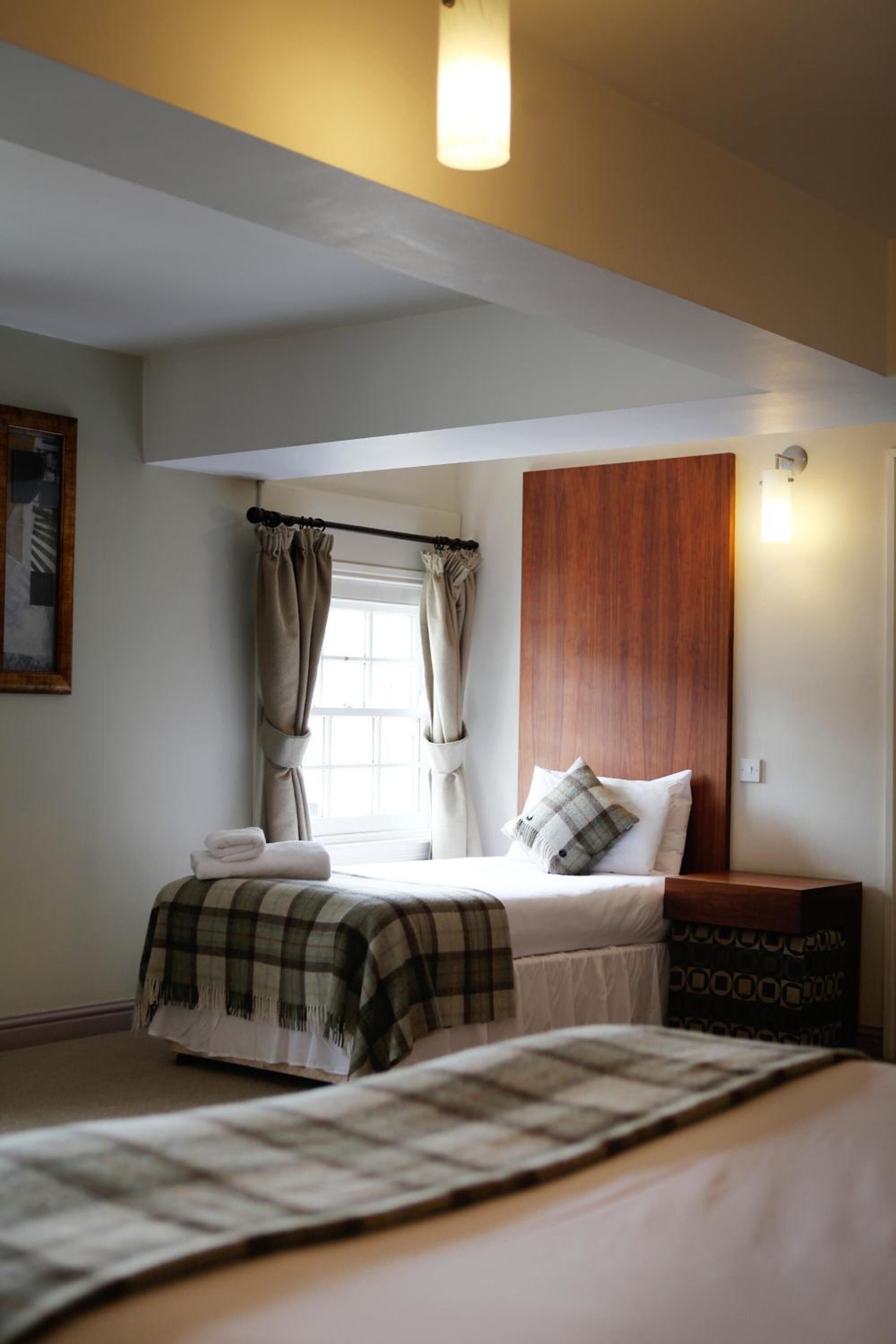 A bedroom with a bed, a bedside table, a window with curtains, a headboard, and a wall-mounted light fixture.