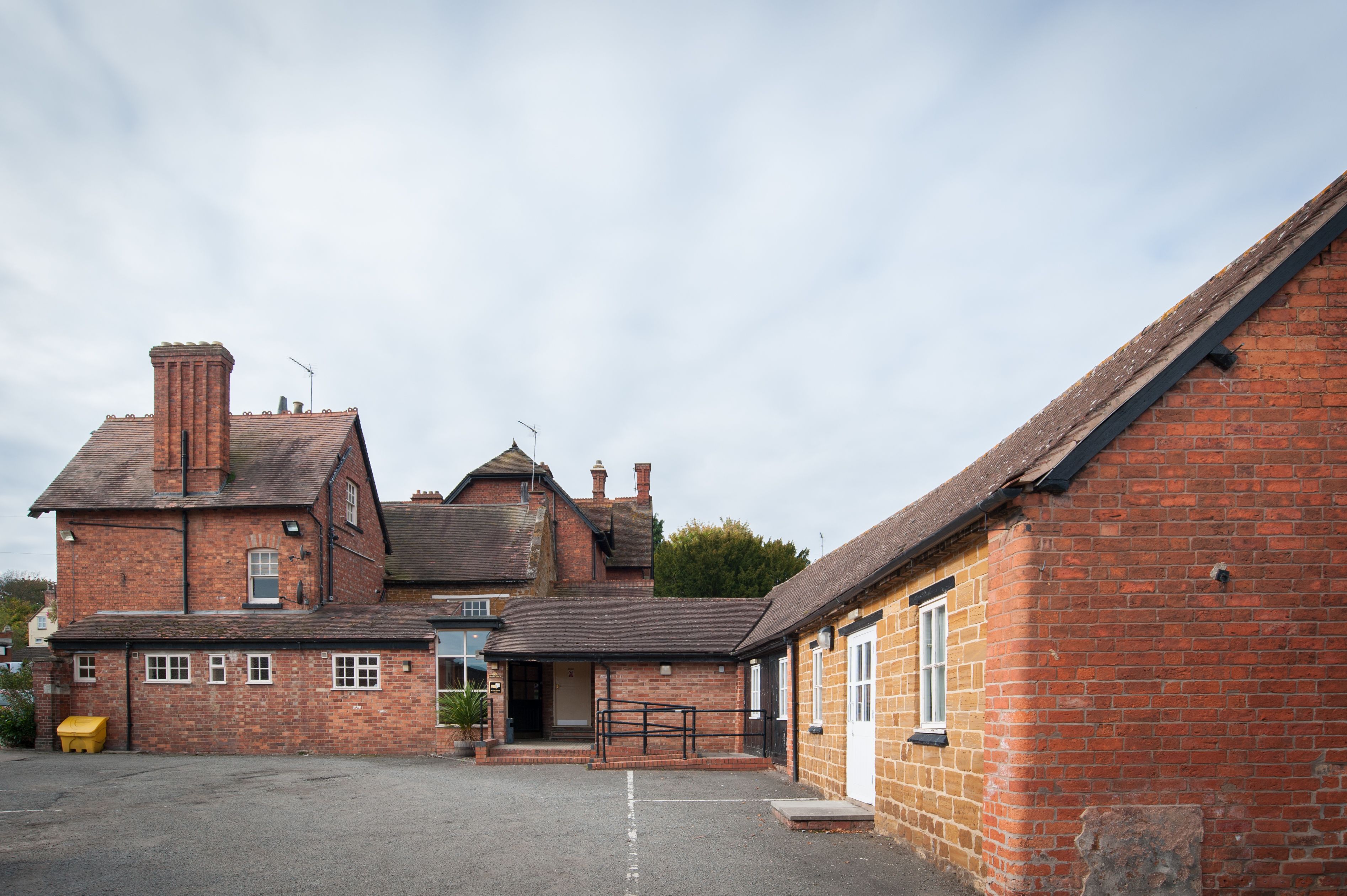 A traditional brick building with a large central section and smaller wings, featuring a gravel driveway and a small entrance gate.